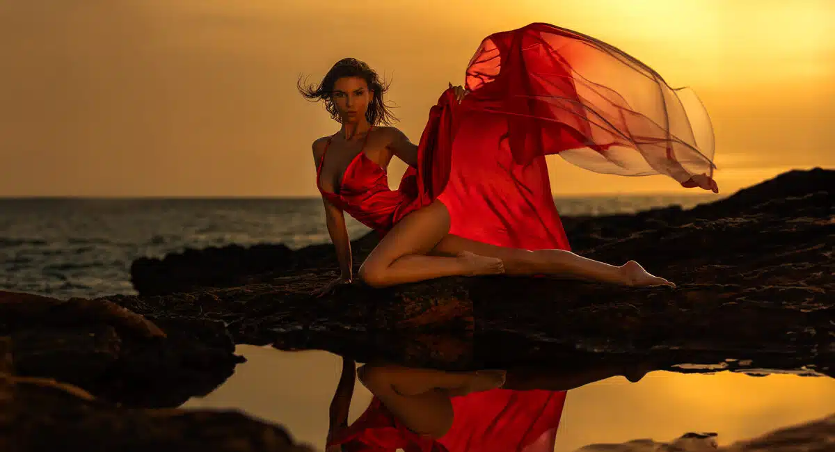 Marcus Boéll Photographer Mallorca Fotomodel in einem fließenden roten Kleid posiert auf einem Felsen am Meer bei Sonnenuntergang, mit einem Teil des Kleides, das im Wind weht.